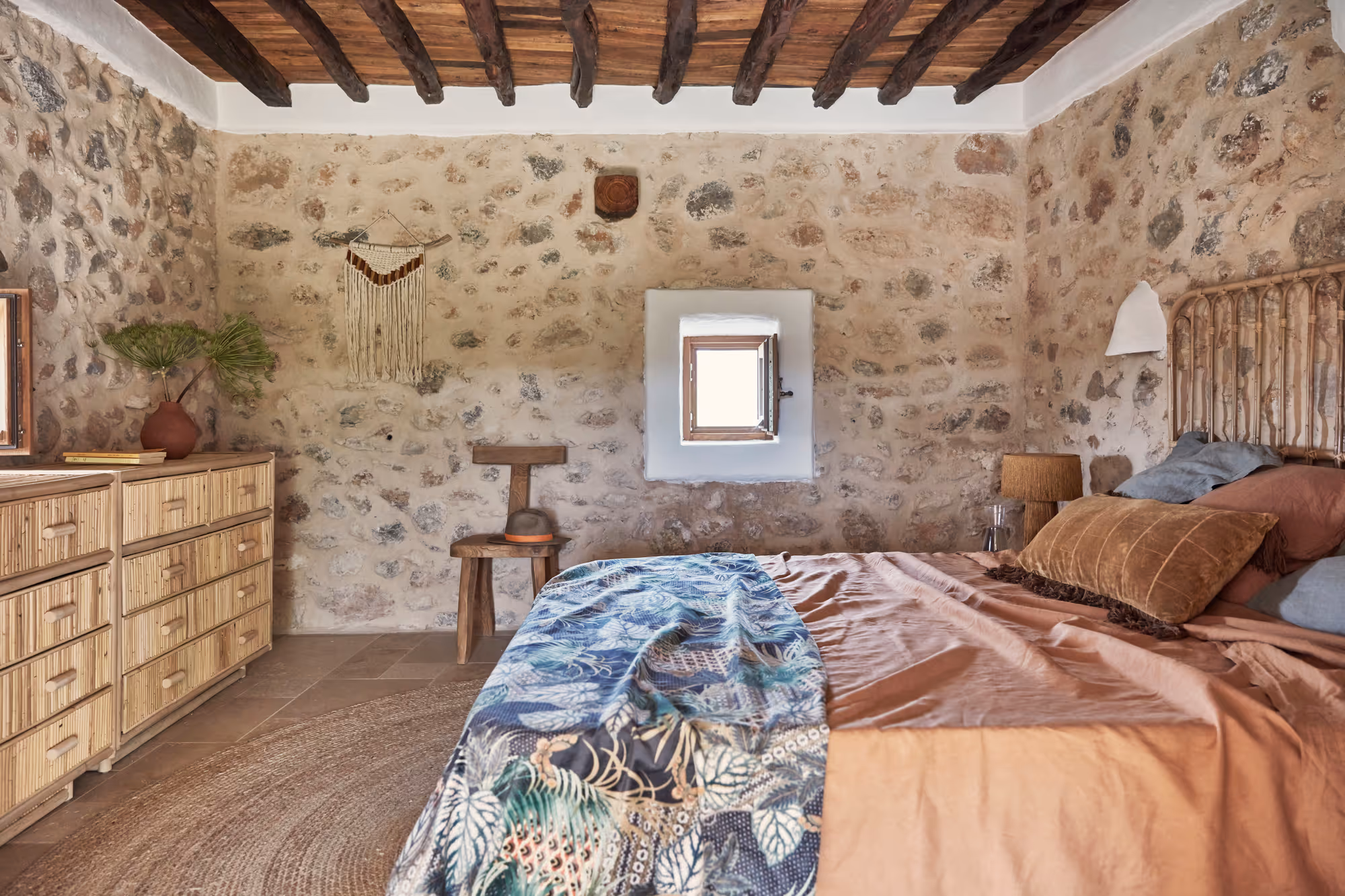 Chambre rustique avec murs en pierre, poutres apparentes, lit recouvert d'une literie à motifs feuillages marron et bleu, commode en bois et petite fenêtre.