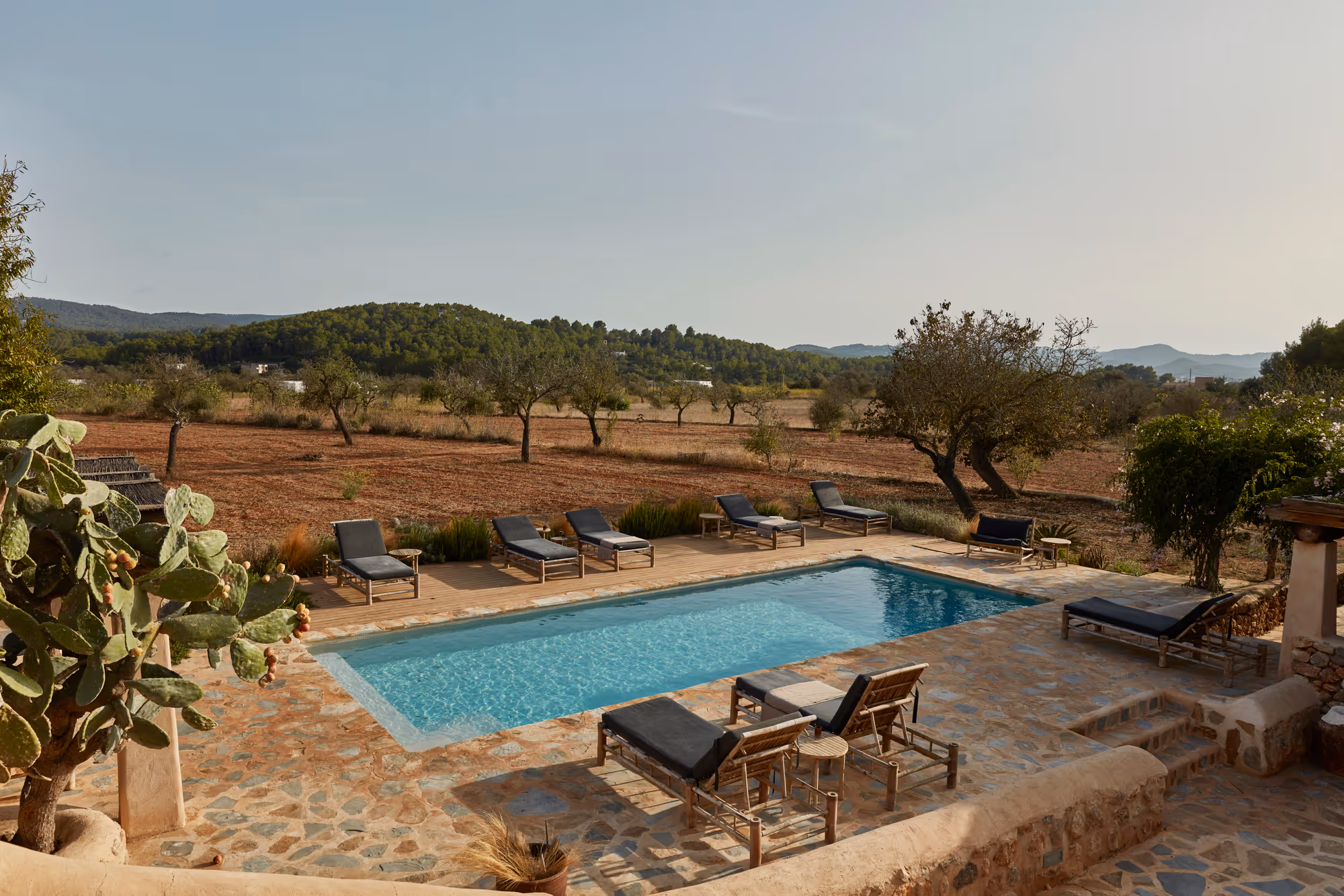 Piscine rectangulaire extérieure entourée d'une terrasse en pierre et de chaises longues, avec un champ sec et des collines en arrière-plan.